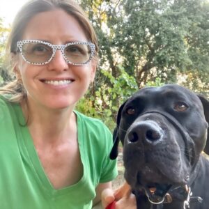 White woman around 50 years of age, with red hair and white glasses, wearing a green shirt with a big black dog.
